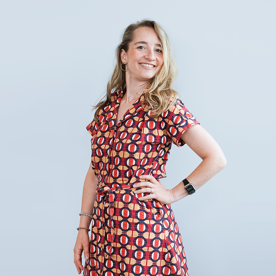  A woman with wavy, shoulder-length hair wears a patterned dress in shades of red and orange. She smiles and poses confidently against a light blue background.